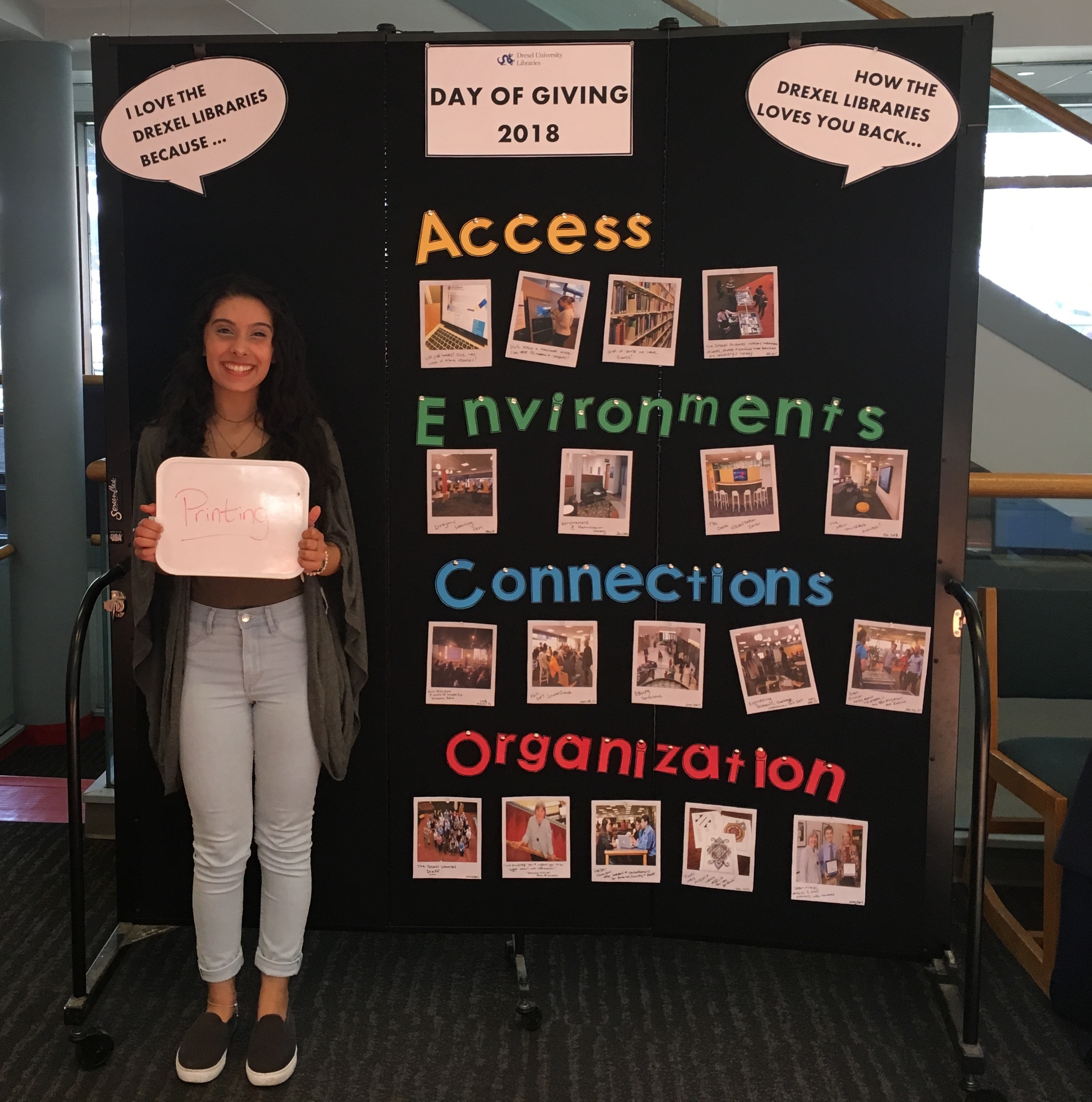 A woman holds a small white dry erase board with writing on it. She is standing in front of a large board with photos on it and the words Access, Environments, Connections, Organization.